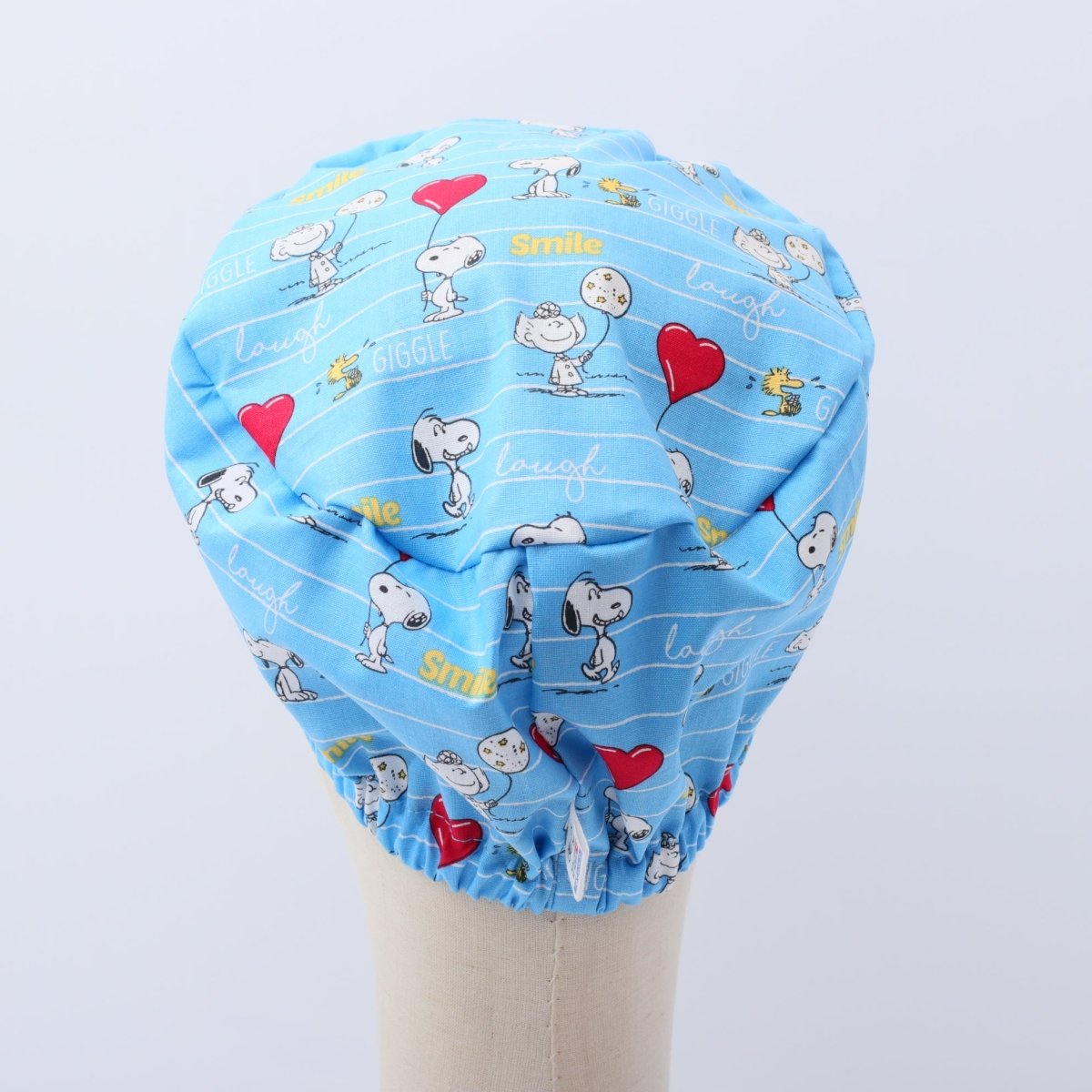 Top view of a blue bouffant scrub cap featuring Snoopy cartoon characters, red hearts and words like smile, laugh and giggle; custom handmade by Medicus Caps.