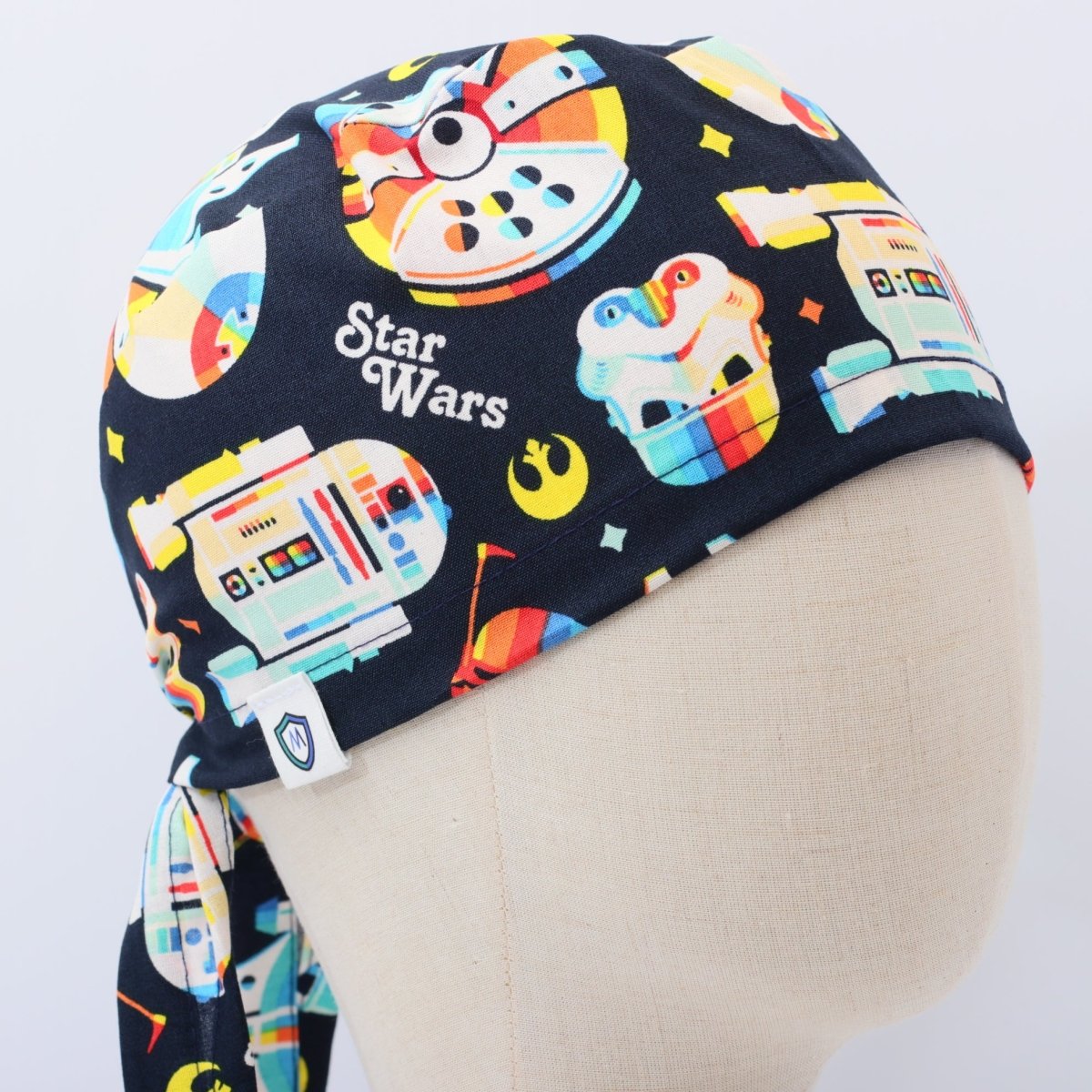 Custom dark blue scrub cap by Medicus Caps featuring colorful space droid robots and galactic motif print inspired by science fiction; side view on mannequin with tie straps.