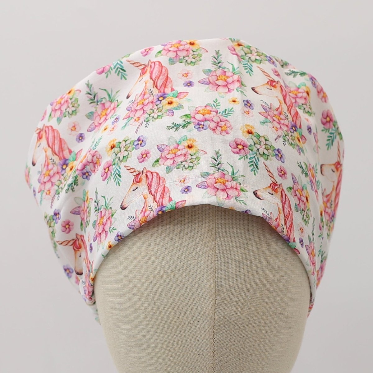 Bouffant style white custom scrub cap featuring pink unicorns and colorful floral patterns. Handmade unicorn floral scrub cap by Medicus Caps with roomy fit ideal for long hair or bouffant style.