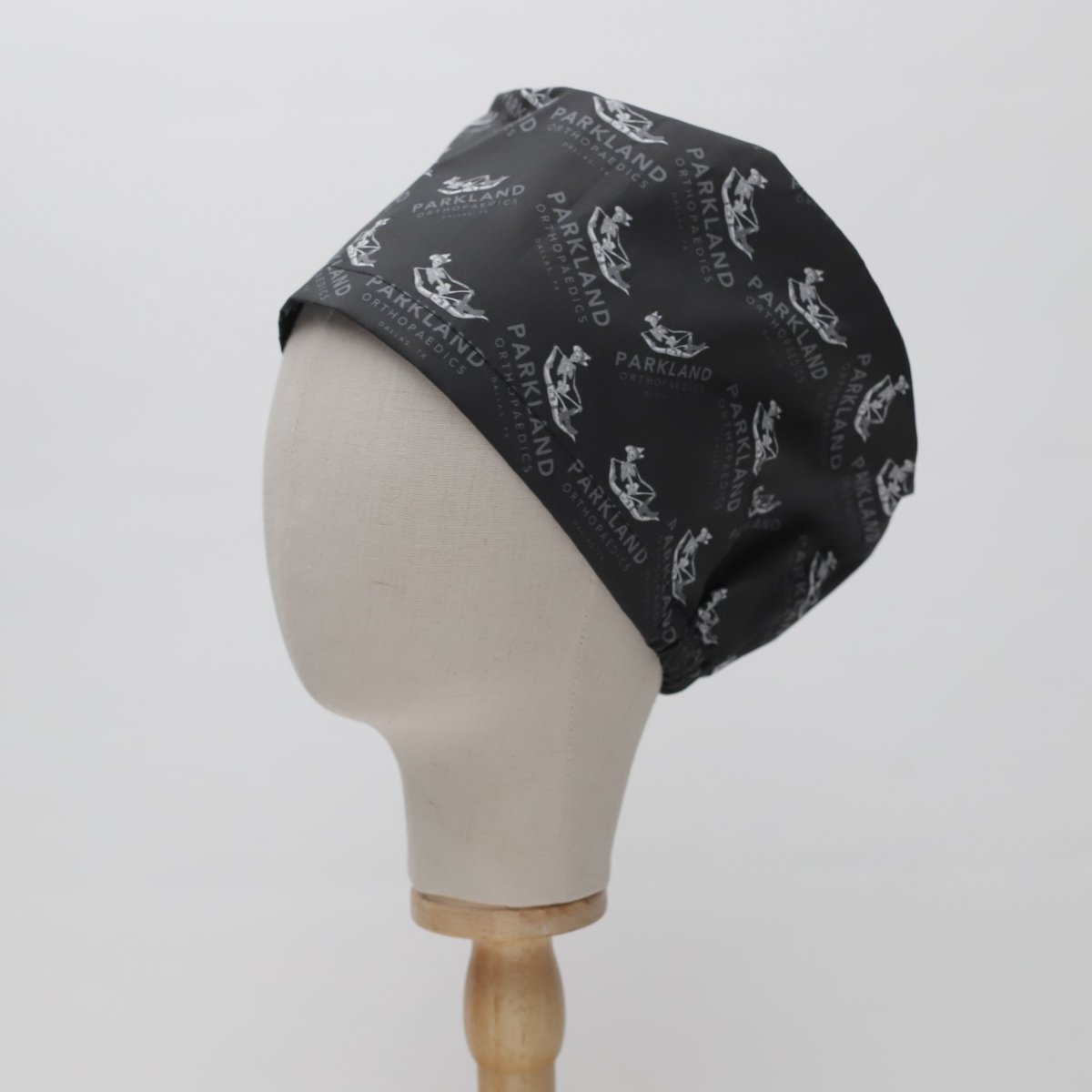 The All Over Custom Printed Fabric Scrub Cap by Medicus Scrub Caps is shown on a mannequin head. It features a black cloth design with white text and logos using licensed fabric against a plain white background.