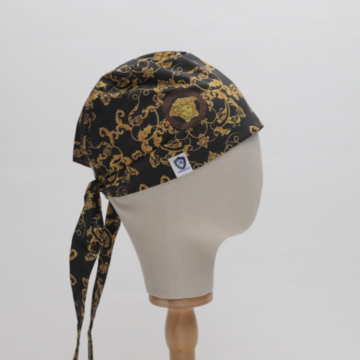 A mannequin head features an All Over Custom Printed Fabric Scrub Cap by Medicus Scrub Caps, displaying a black bandana with gold ornate patterns and a central medallion design, similar to licensed fabric art. Its tied at the back with a side tag visible.