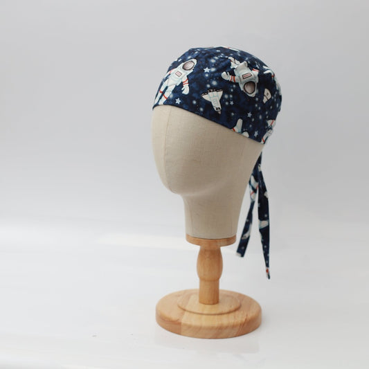 A mannequin head displays the Astronauts Spaceman Scrub Cap by Medicus Scrub Caps, featuring astronauts and celestial prints. Its mounted on a wooden stand against a plain white backdrop.