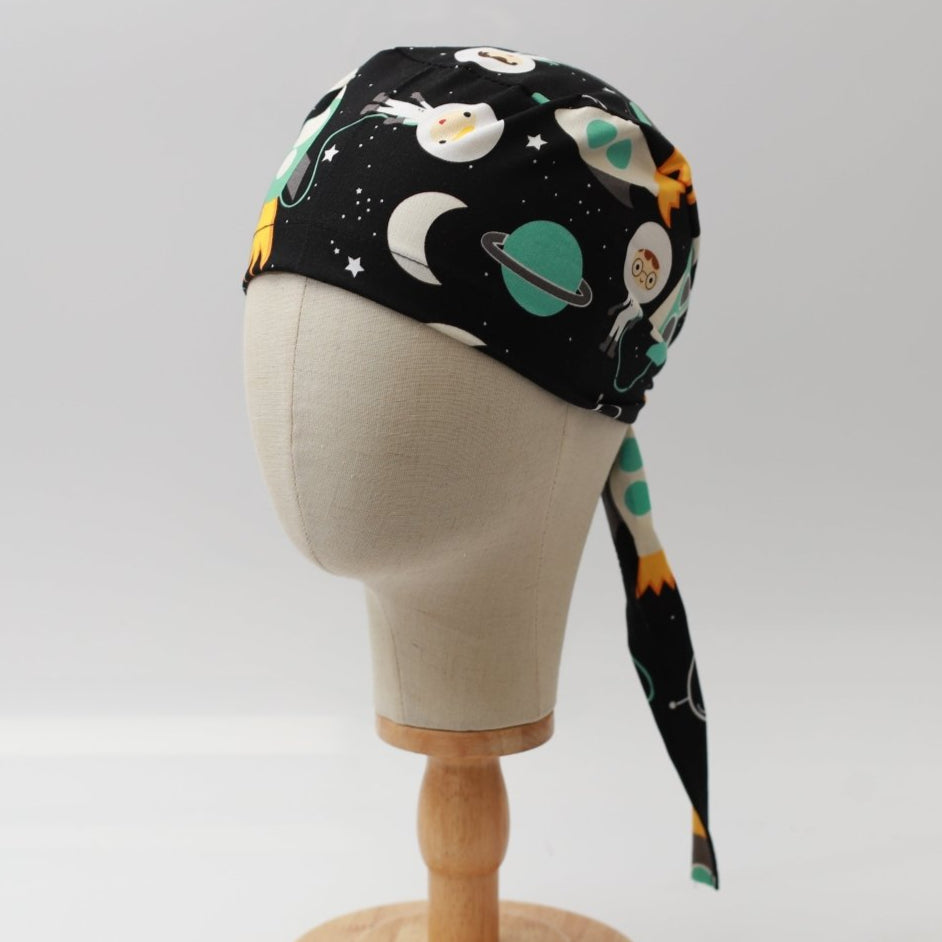 Mannequin head wearing Medicus Scrub Cap with cartoon spaceships blue print, on a wooden stand against a white backdrop.