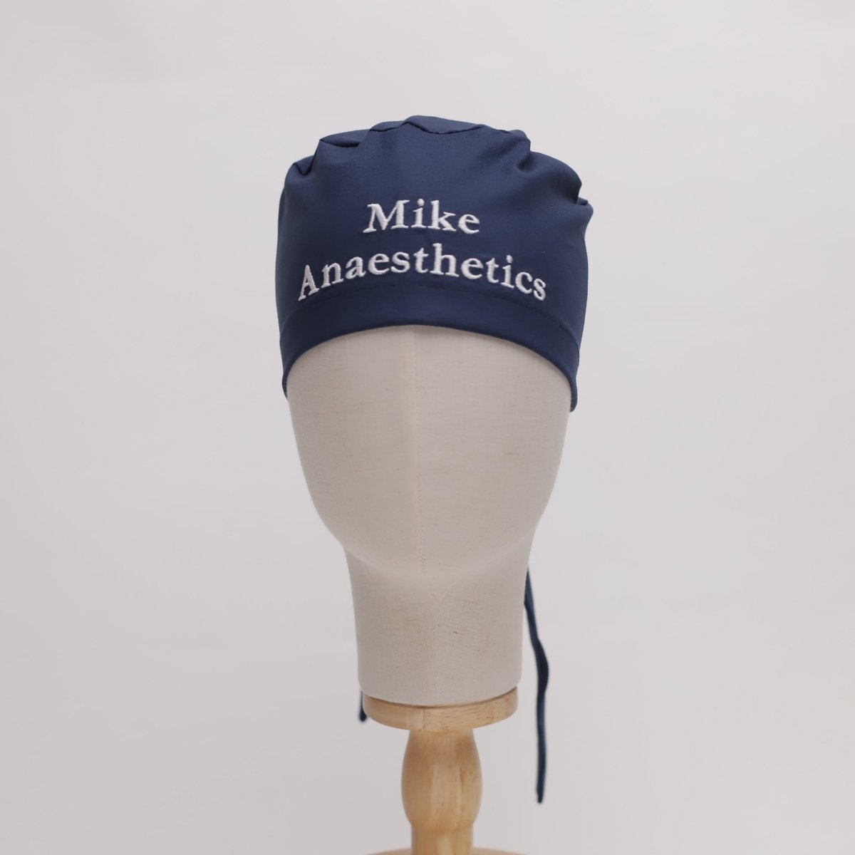 A mannequin head displays a navy cap from Medicus Scrub Caps, featuring high-quality fabric with Mike Anaesthetics embroidered in white, emphasizing its custom design against the plain white background.