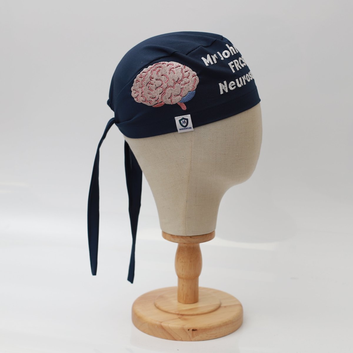 A mannequin head displays a Medicus Scrub Caps dark blue surgical cap, featuring high-quality fabric with pink brain embroidery and Mr John FRCS Neurosurgery text. The custom scrub cap sits elegantly on a wooden stand against a plain white background.