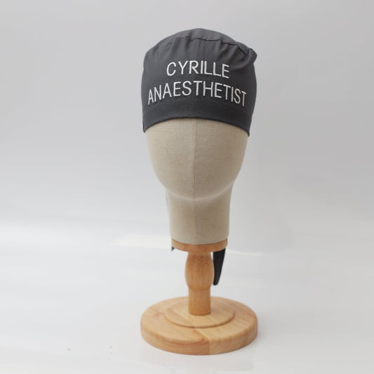 A mannequin head features a black Medicus Scrub Caps custom surgical cap with CYRILLE ANAESTHETIST in white text. It elegantly rests on a wooden stand against a pristine white background.