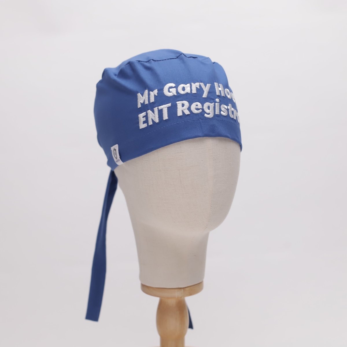 Mannequin head wearing a blue scrub cap with white embroidered text 'Mr Gary Hock ENT Registrar.'