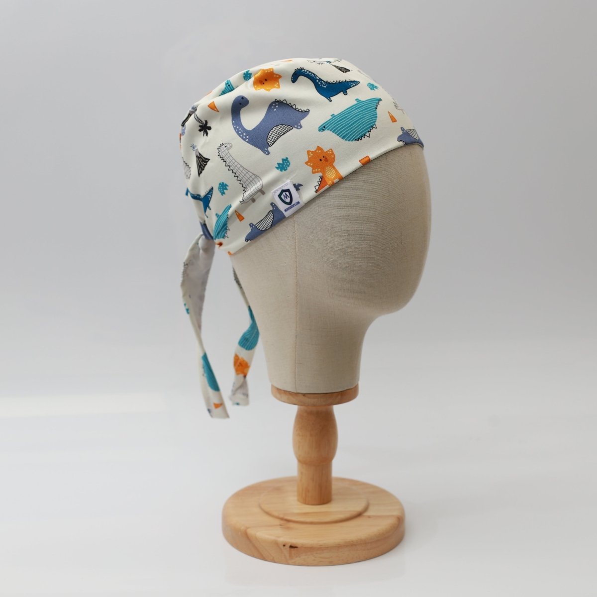 A mannequin head displays the Medicus Scrub Caps Dinosaurs Purple Blue Orange Scrub Cap, adorned with vibrant animal illustrations like whales and dinosaurs. Its designed for healthcare professionals, ties at the back, and is set against a plain white background for a medical field adventure.