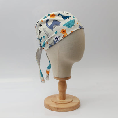A mannequin head displays the Medicus Scrub Caps Dinosaurs Purple Blue Orange Scrub Cap, adorned with vibrant animal illustrations like whales and dinosaurs. Its designed for healthcare professionals, ties at the back, and is set against a plain white background for a medical field adventure.