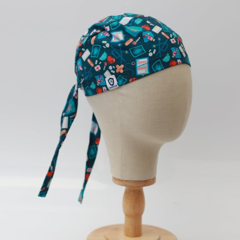 A mannequin head sports a Medicus Scrub Caps navy scrub cap, adorned with medical symbols such as syringes, band-aids, and pills. Tied securely on a wooden stand against a plain white background, its ideal for dedicated healthcare professionals.