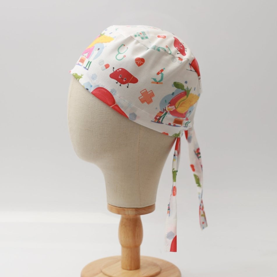 A mannequin head displays a Medicus Scrub Caps Human Body Gall Bladder Liver Pancreas Scrub Cap with colorful medical-themed illustrations, tied at the back, on a wooden stand.