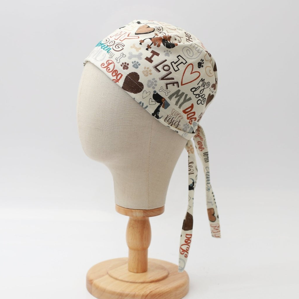 A mannequin head displays the I Love My Dogs scrub cap by Medicus Scrub Caps, featuring a vibrant dog-themed design with bones, paw prints, and phrases. Ideal for veterinary professionals, it is reusable with two long ties at the back and is presented on a wooden stand.