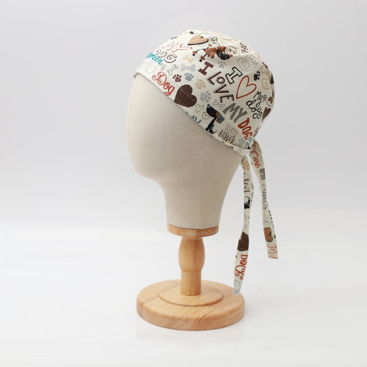 A mannequin head displays the I Love My Dogs scrub cap by Medicus Scrub Caps, featuring a vibrant dog-themed design with bones, paw prints, and phrases. Ideal for veterinary professionals, it is reusable with two long ties at the back and is presented on a wooden stand.