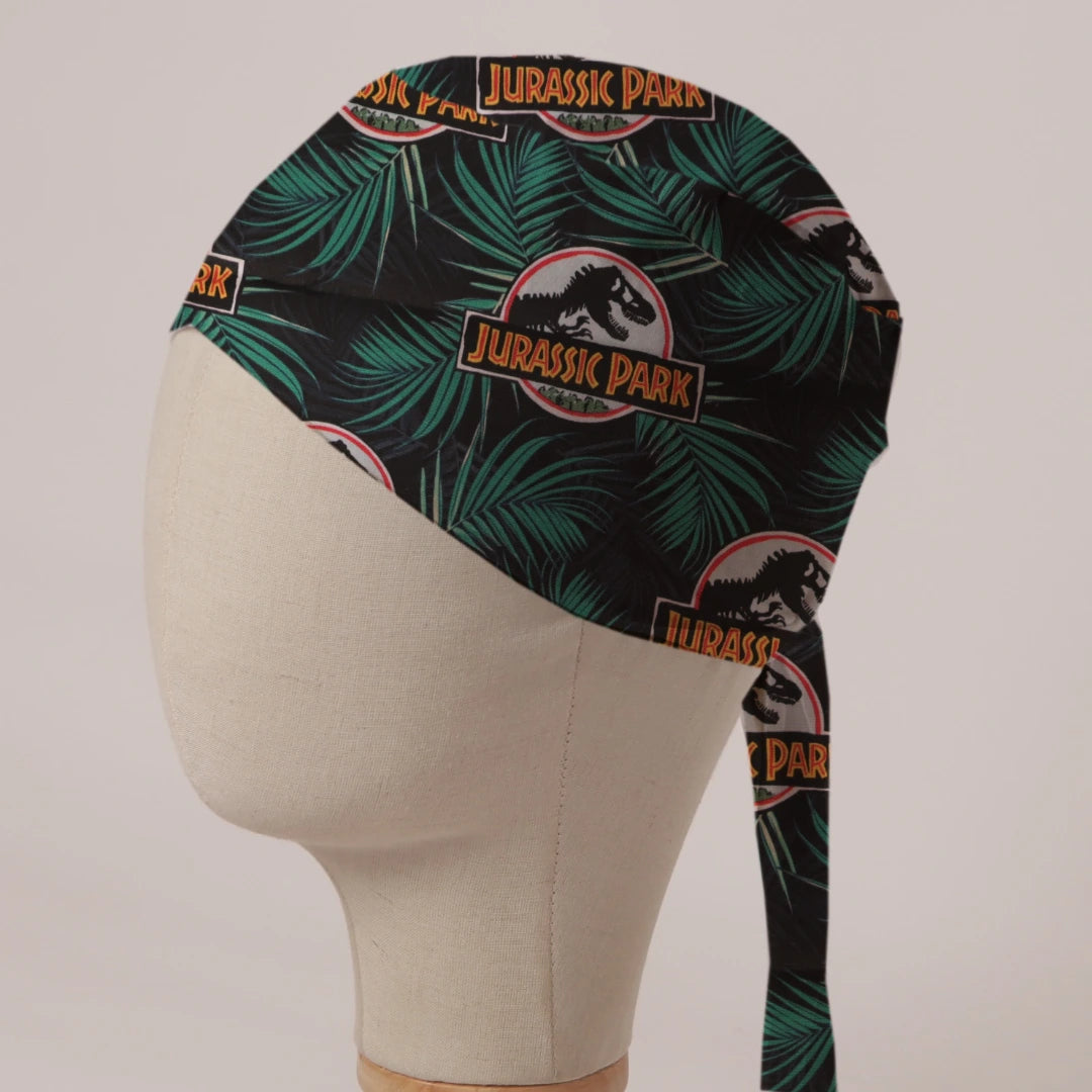 Jurassic Park Logo Jungle fabric scrub cap in Bandana tie-back style: Jurassic Park logo among palm leaves on black