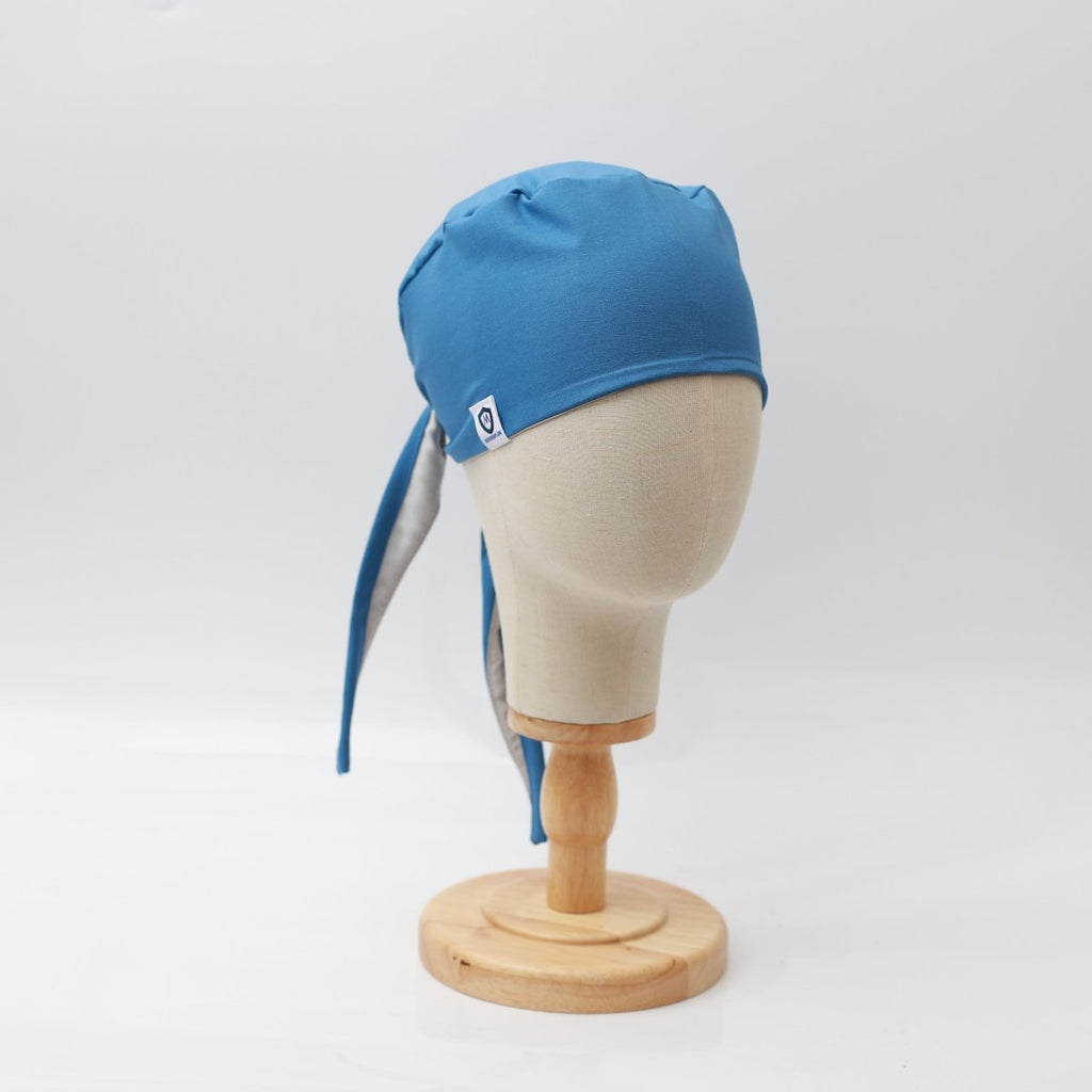 A blue Plain Satin Lined Scrub Cap by Medicus Scrub Caps is elegantly displayed on a beige mannequin head with a wooden stand. The eco-friendly cap, featuring back ties and a logo tag, stands out against the plain white background.