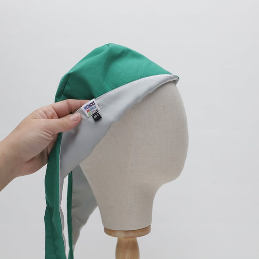 A hand holds a sleek green and white scrub cap on a mannequin head, crafted with sustainability in mind. A tag reads Medicus Scrub Caps. This eco-friendly, satin-lined piece is a stylish nod to simplicity against a plain white backdrop.