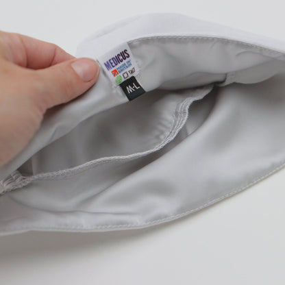 A person holds the edge of a white Medicus Scrub Cap, showing labels with laundry symbols and MEDICUS M-L. The stitching and texture indicate quality. This reusable Plain Satin Lined Scrub Cap, available in 22 colors, provides eco-friendly care and comfort.