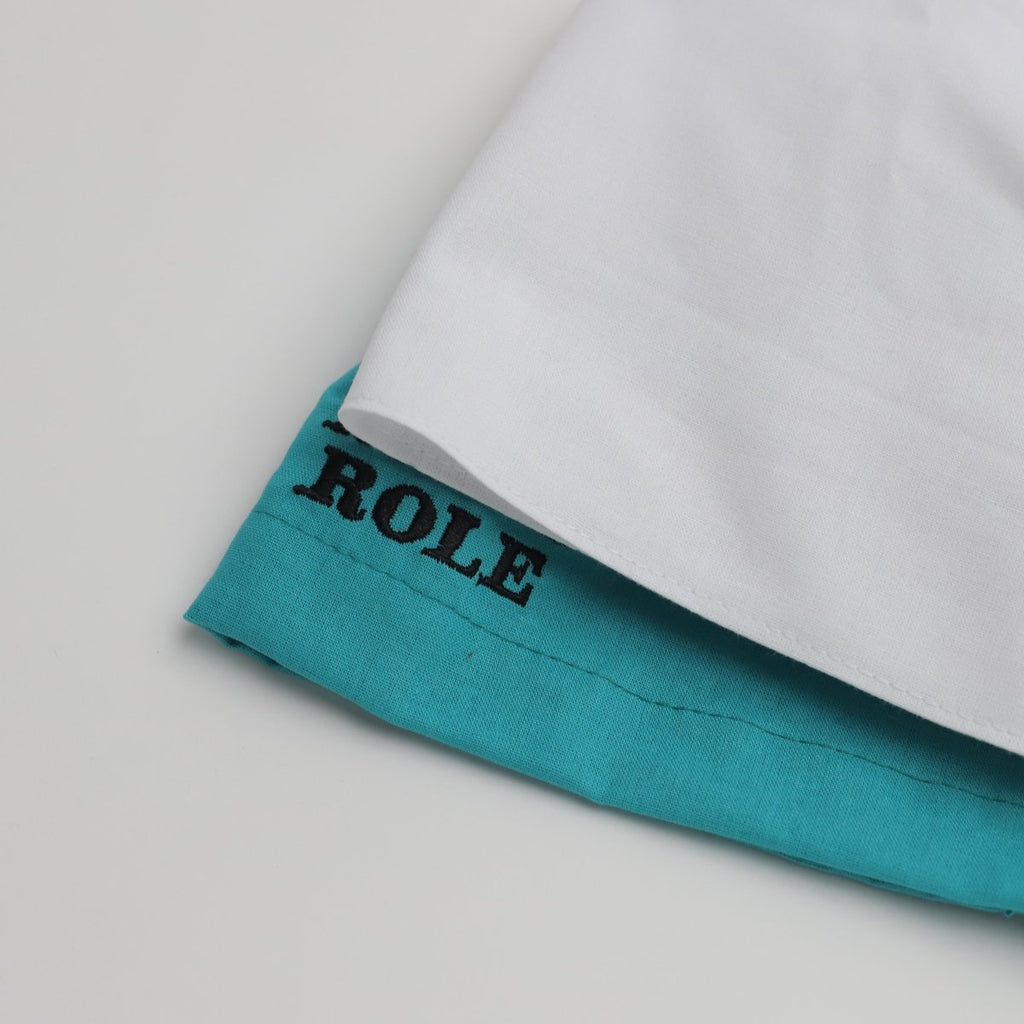 A Medicus Scrub Caps product—a Plain Satin Lined Scrub Cap in white and teal—sits folded on a light surface. The teal part displays partial black text reading ROLE, showcasing its eco-friendly design among 22 available colors.