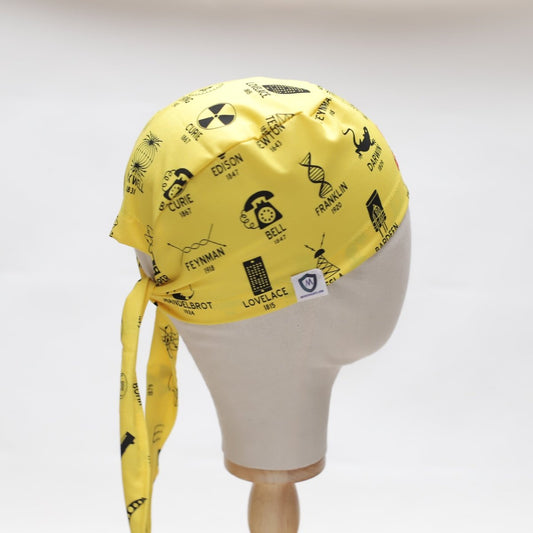 The Science Founders Scrub Cap by Medicus Scrub Caps is a yellow bandana with black scientific symbols and names like Edison and Franklin. Displayed on a mannequin head, it features a tied section at the back and a small logo tag, blending style with sustainability.