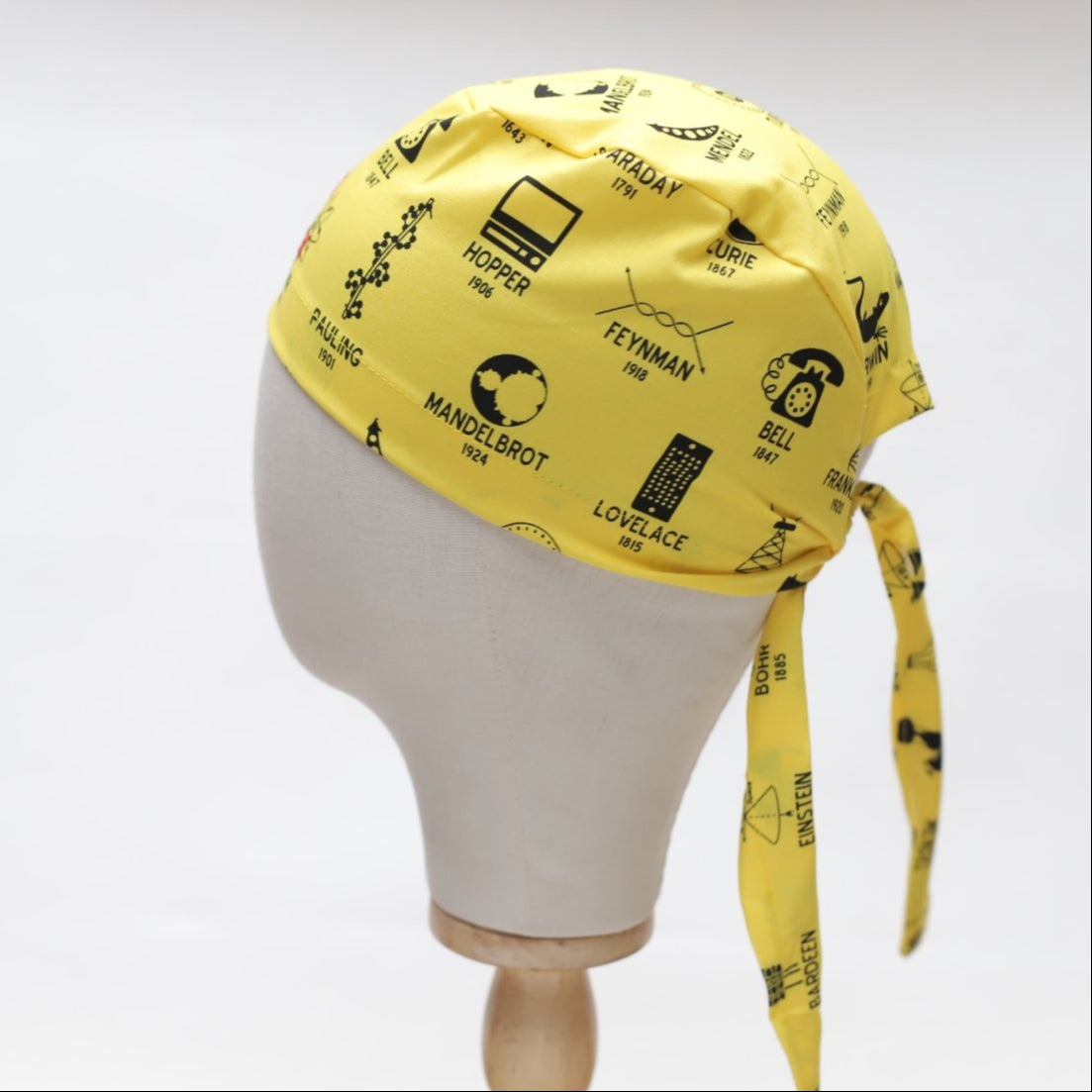 A mannequin head displays a yellow Science Founders Scrub Cap by Medicus Scrub Caps, featuring black graphics and names of iconic scientists such as Hopper and Feynman, tied at the back against a white backdrop.