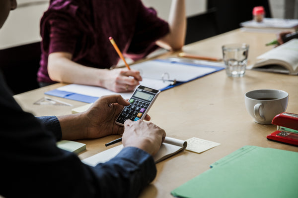 People work at a desk covered with documents and stationery. One person uses a calculator, and another takes notes. A coffee mug, stapler, and water glass are on the table. The scene conveys a collaborative work environment.