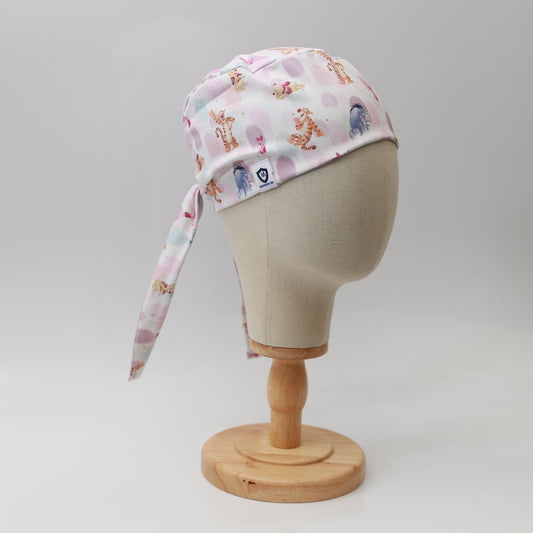 A mannequin head displays a custom Medicus Scrub Cap named Winnie The Pooh + Friends Finger Paint, featuring playful animal illustrations like tigers and pink stars. The tie-back cap rests on a wooden stand against a white background, merging charm with sustainable healthcare.