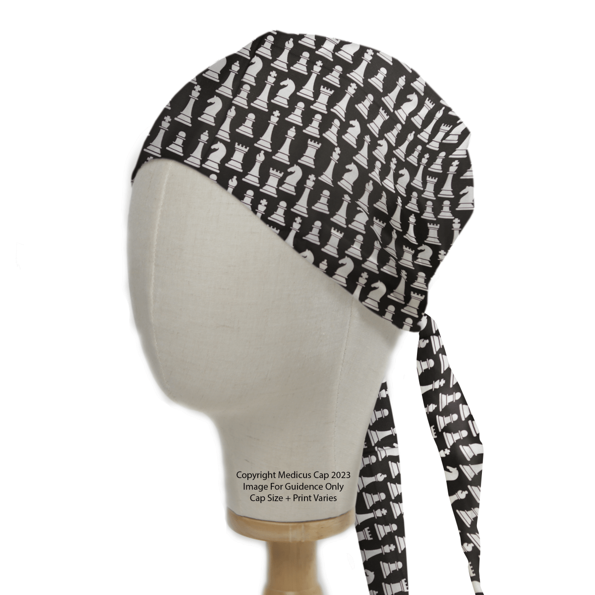 A mannequin head displays the Chess Pieces Classic Black + White Scrub Cap, featuring white chess pieces symbolizing strategic brilliance. The black cap has extended ties at the back. The image mentions Medicus Scrub Caps 2023 copyright, with notes on image guidance and size variance.