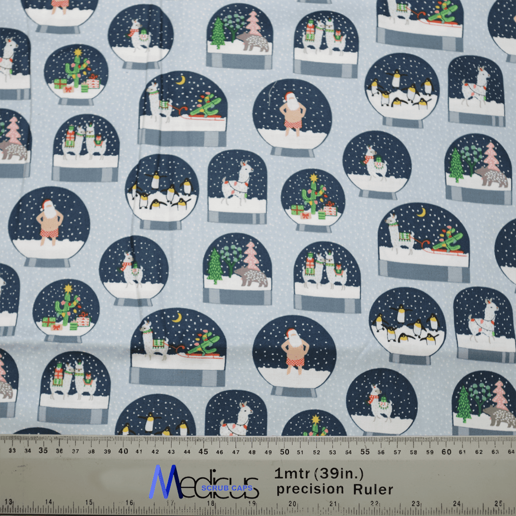 The Christmas Snow Globes Blue Scrub Cap by Medicus Scrub Caps features a winter-themed design with cartoon snow globes showcasing holiday scenes like snowmen, penguins, and Christmas trees. A ruler at the bottom shows measurements in inches and meters.