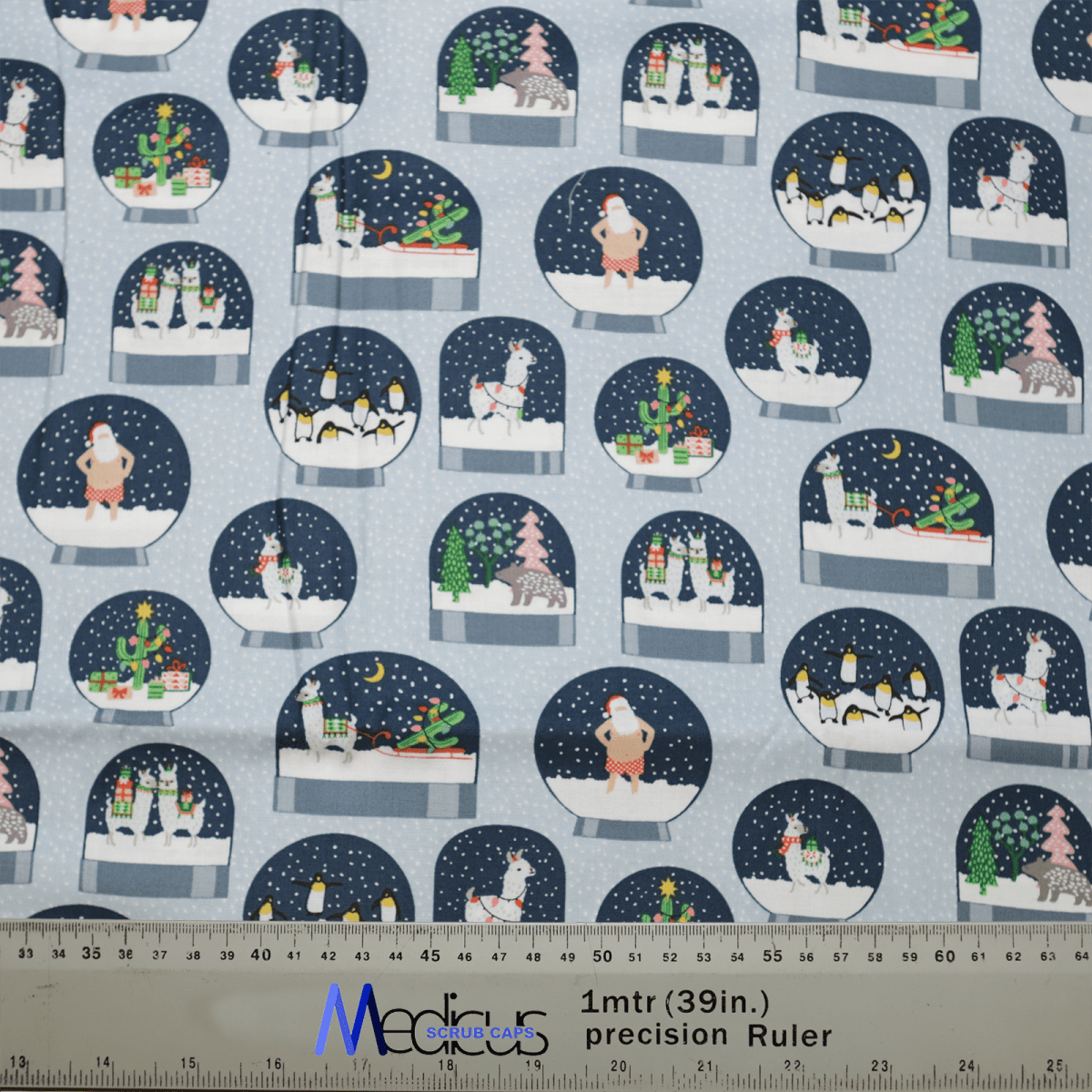 The Christmas Snow Globes Blue Scrub Cap by Medicus Scrub Caps features a winter-themed design with cartoon snow globes showcasing holiday scenes like snowmen, penguins, and Christmas trees. A ruler at the bottom shows measurements in inches and meters.