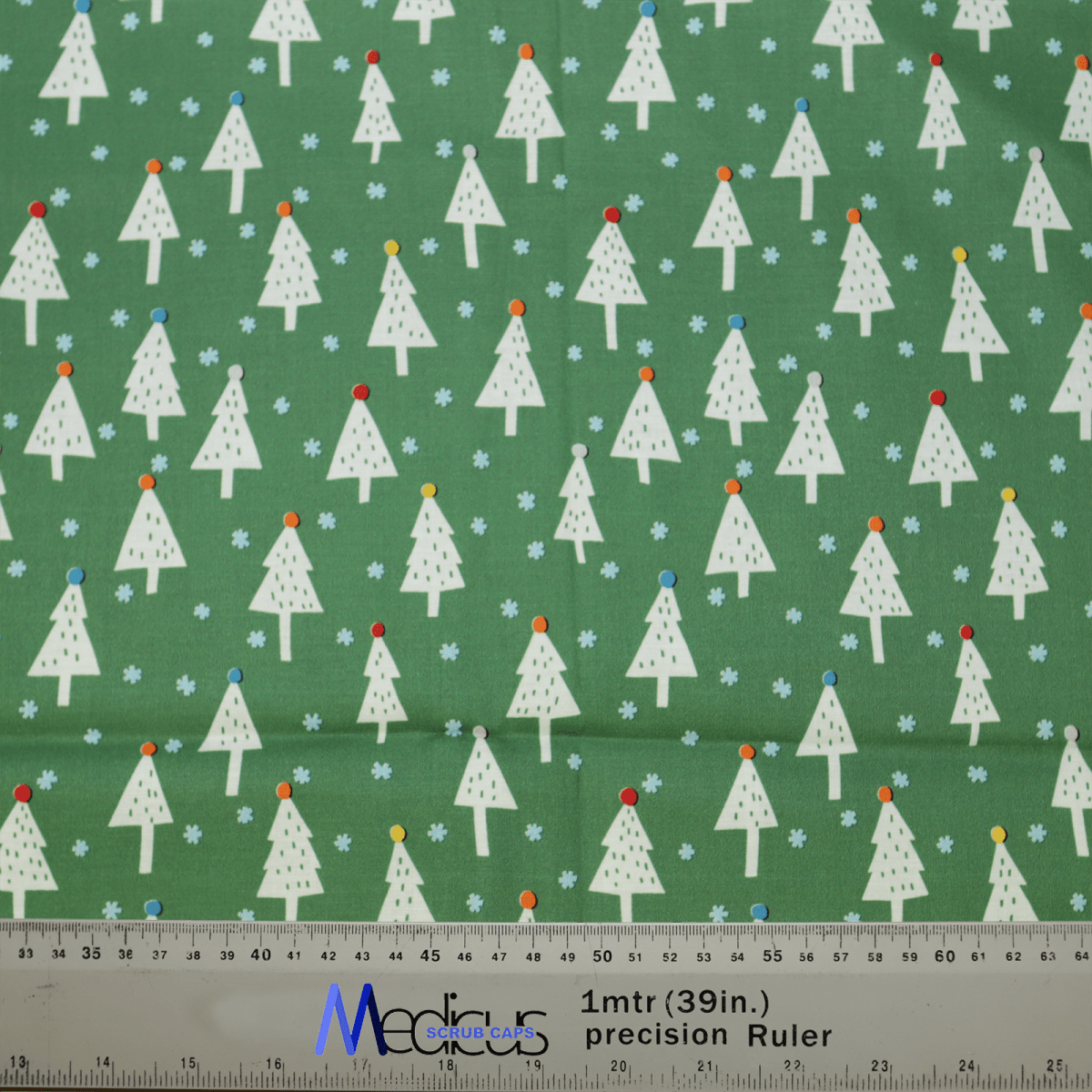This Medicus Scrub Caps product, the Christmas Trees Scrub Cap, features green fabric with stylized white Christmas trees topped with colorful dots. Small blue flowers enhance its charm, and a ruler at the bottom offers precise fit measurements in both inches and centimeters.