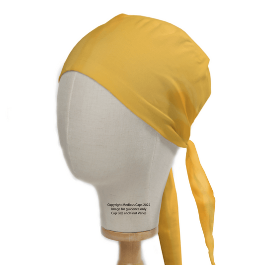 A mannequin head displays a Medicus Scrub Cap styled like a bandana, tied at the back. The label identifies it as a 2022 Classic Plain Gold Yellow Scrub Cap | Theatre Hat, praised for its exceptional durability.