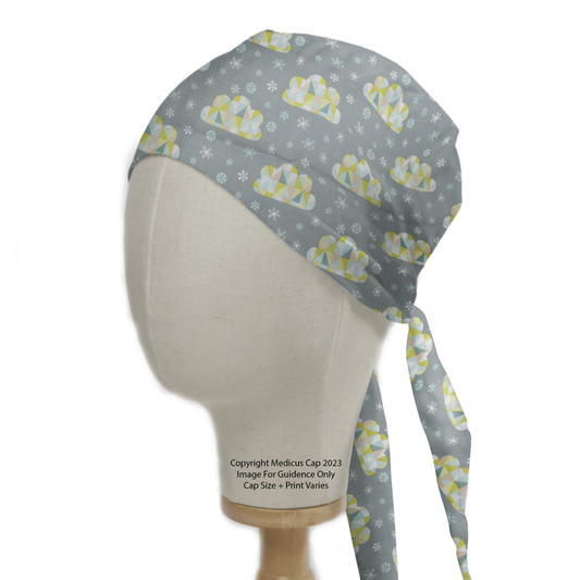 A mannequin head models the Clouds Abstract Stars Scrub Cap by Medicus Scrub Caps, featuring gray with colorful geometric bears and white snowflakes. The cap has long ties at the back and is elegantly displayed on a wooden stand.