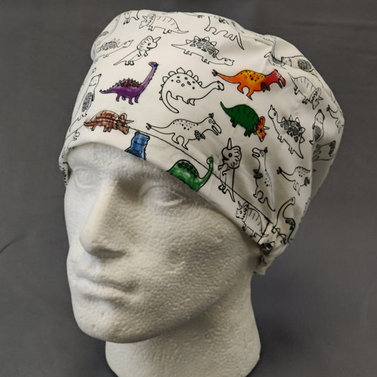 A white mannequin head displays a Medicus Scrub Caps Colour Me In scrub cap, featuring colorful dinosaur patterns in green, orange, red, and blue on a white background for a playful design.