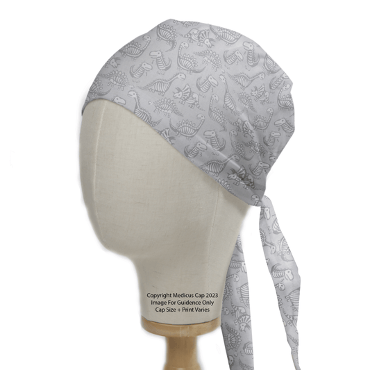 The mannequin head displays the Dinosaur Dig #2 Scrub Cap by Medicus Scrub Caps in light gray, featuring T-Rex, triceratops, and stegosaurus patterns. It includes long ties at the back, symbolizing the inclusive spirit of healthcare heroes.