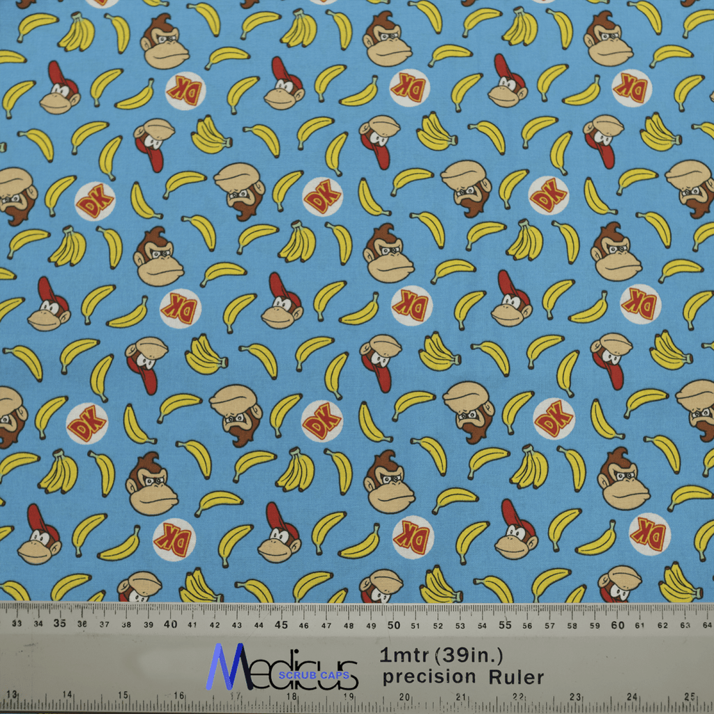 The DK Donkey Kong Bananas Blue Scrub Cap by Medicus Scrub Caps features playful gorilla characters, bananas, and DK logos on light blue fabric with measurement markings.