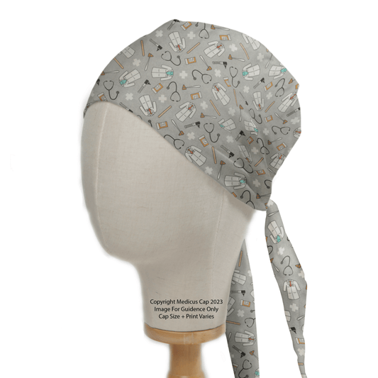 The Medicus Scrub Caps Doctor GP General Grey Scrub Cap, displayed on a wooden stand, features a medical icons pattern of stethoscopes, pills, bandages, and hearts. It is reusable and hypoallergenic, designed to tie neatly at the back for a secure fit.