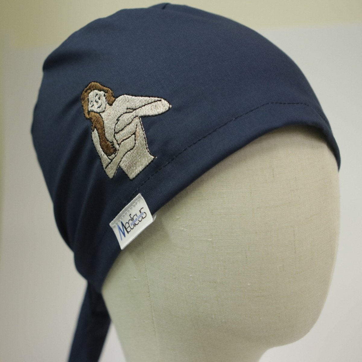 A mannequin head showcases the Embroidery - Breast Cancer Awareness - Scrub Cap from Medicus Scrub Caps, featuring an embroidered person with a cat, a reusable design promoting breast cancer awareness, and a tag reading Medicus.