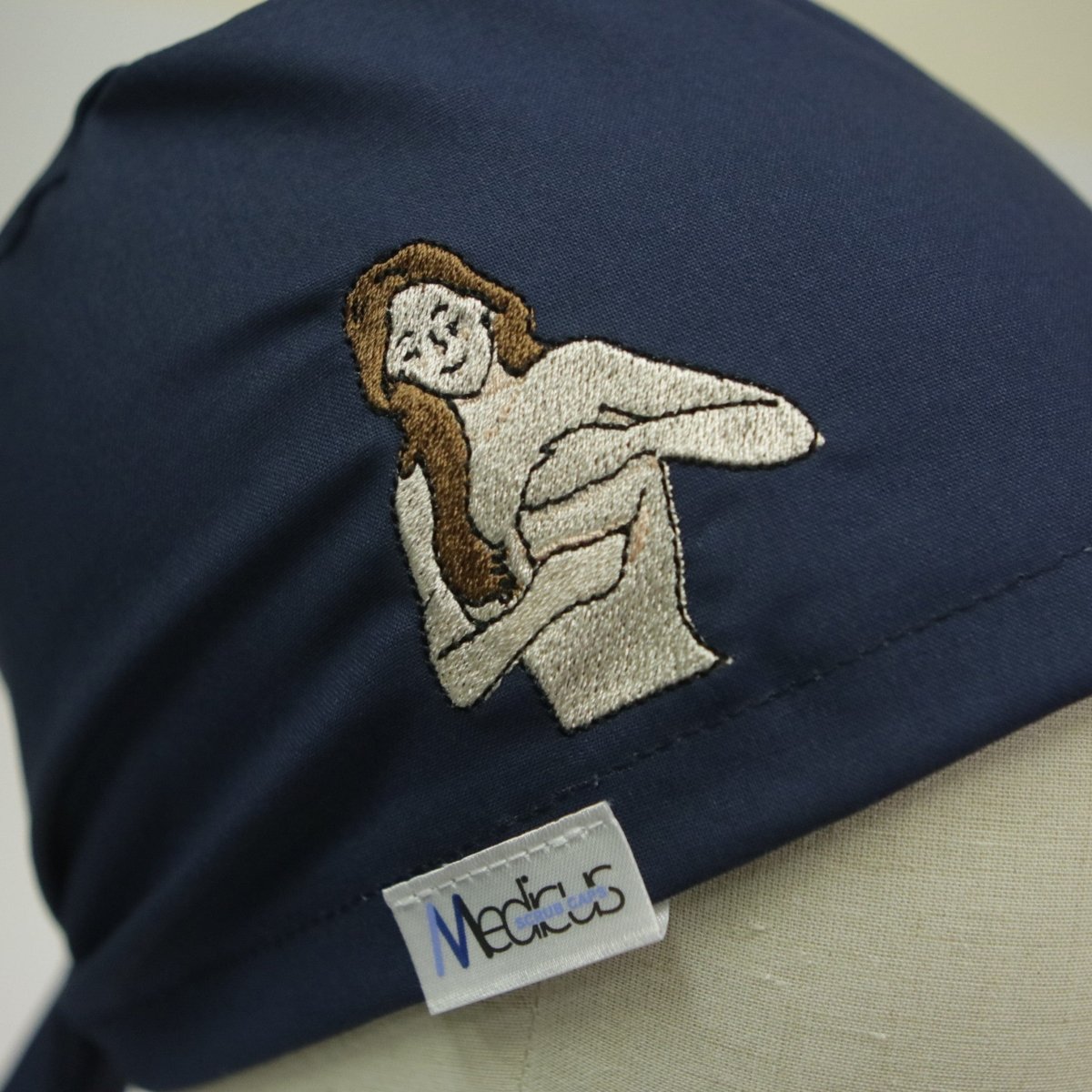 The blue scrub cap, embroidered with a smiling person with long hair performing a medical exam gesture, subtly supports breast cancer awareness. A white label reads Medicus in blue. Perfectly reusable, it adds charm to your medical attire. Product: Embroidery - Breast Cancer Awareness - Scrub Cap by Medicus Scrub Caps.