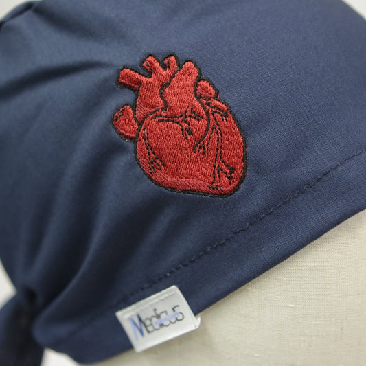 A dark blue scrub cap called Embroidery - Classic Heart from Medicus Scrub Caps showcases a red anatomical heart design with detailed stitching. It features a small bottom label reading Medicus.