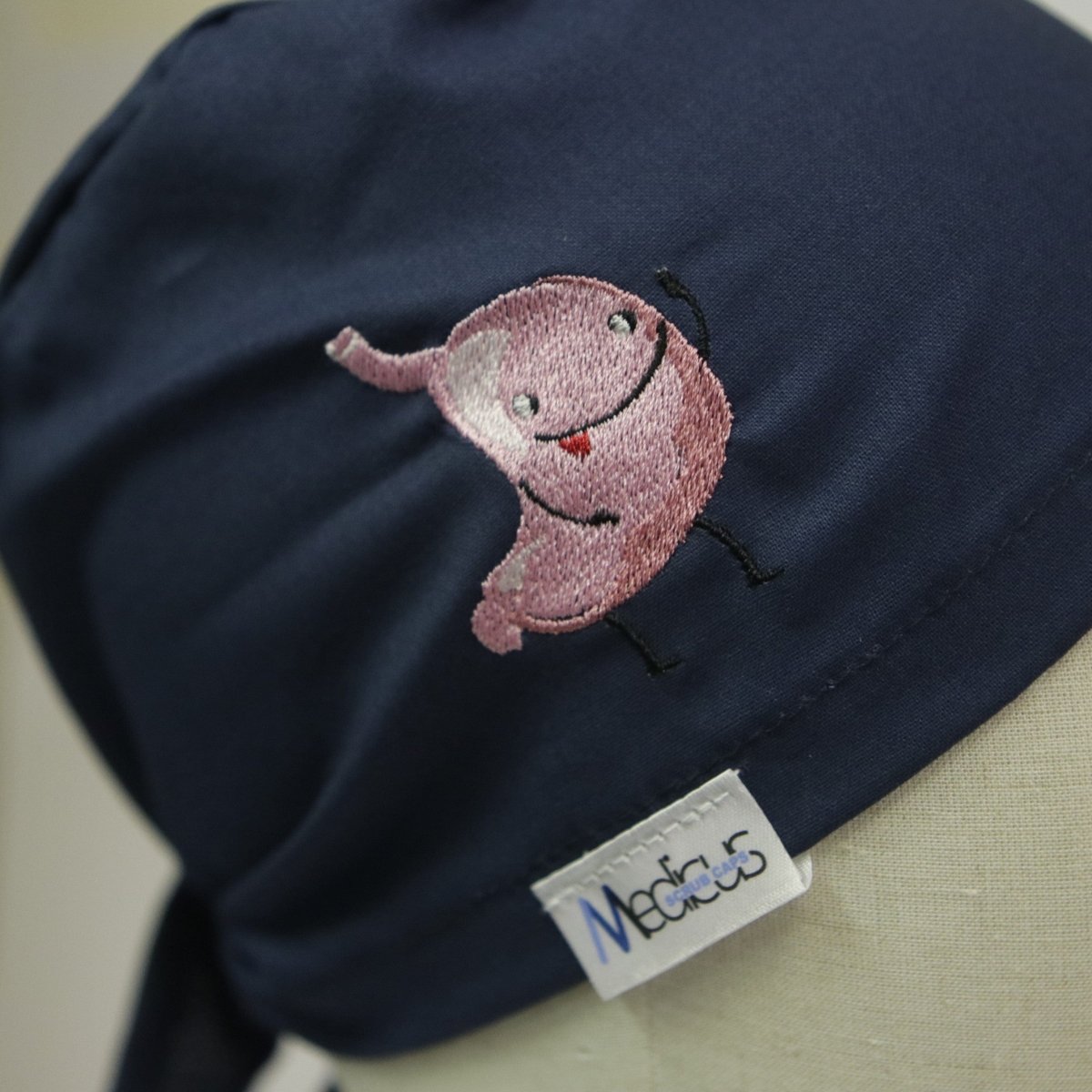 The Medicus Scrub Caps Embroidery - Happy Stomach features a dark blue fabric with an embroidered pink cartoon stomach character that is smiling and waving. A small white tag with Medicus in blue and grey text adds a charming touch, ideal for those seeking embroidered scrub caps.