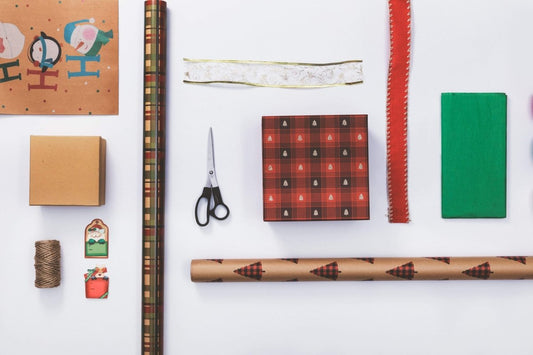 Overhead shot of Medicus Scrub Caps Gift Wrapping supplies on white: wrapping paper, ribbon, scissors, customized twine and tags, a plaid-wrapped box, and a plain box—all arranged for a festive touch.