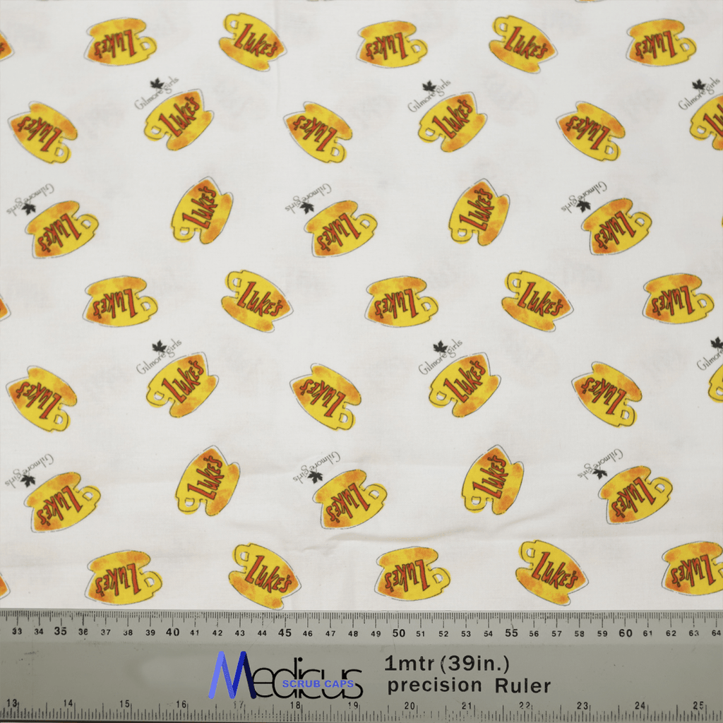 This Medicus Scrub Caps fabric features yellow coffee mugs with Lukes in orange, nodding to Gilmore Girls cafe. Scattered are tiny dog and bird figures. A gray ruler at the bottom shows sizes in inches and centimeters, ideal for crafting your own Gilmore Girls Lukes Cafe Coffee Scrub Cap.