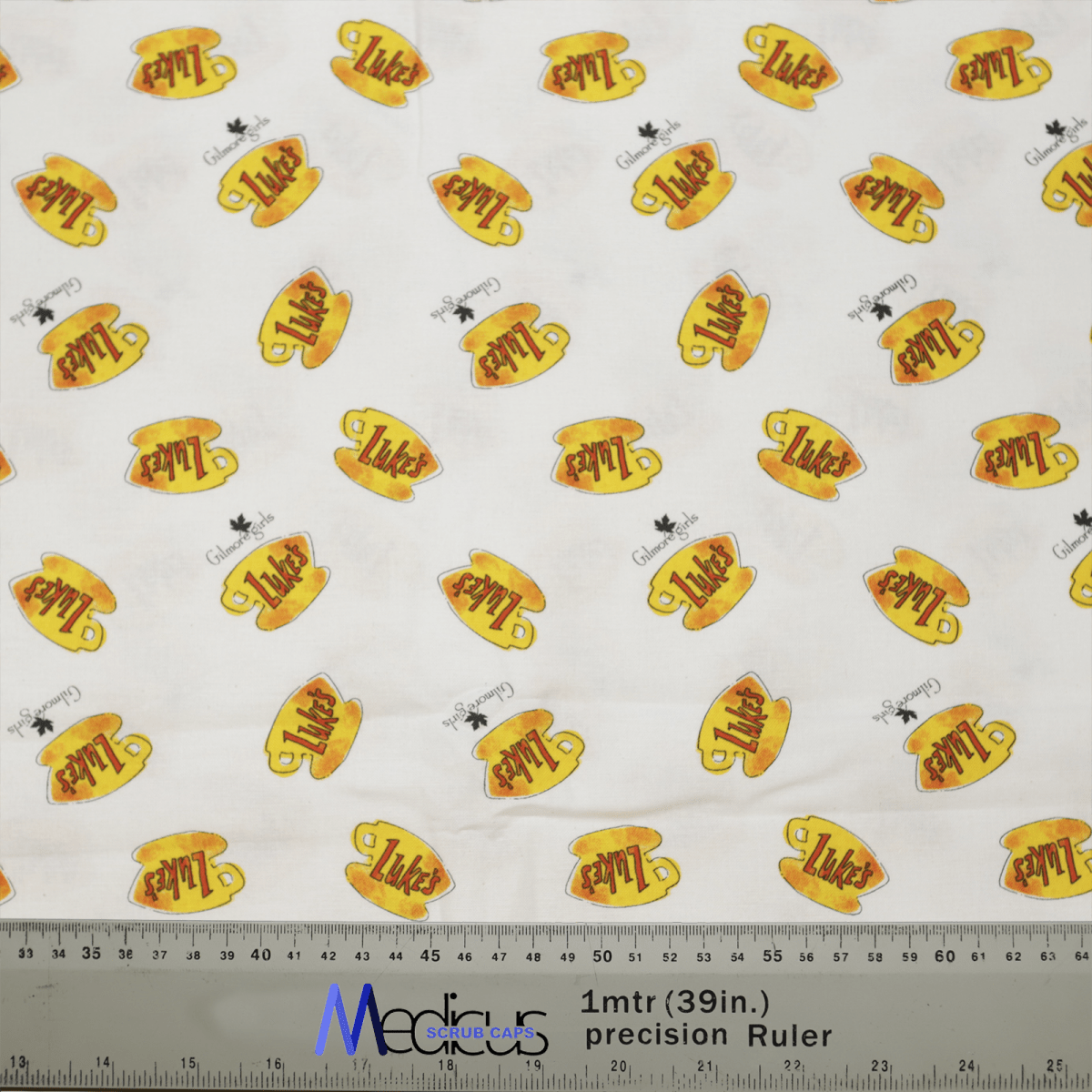 This Medicus Scrub Caps fabric features yellow coffee mugs with Lukes in orange, nodding to Gilmore Girls cafe. Scattered are tiny dog and bird figures. A gray ruler at the bottom shows sizes in inches and centimeters, ideal for crafting your own Gilmore Girls Lukes Cafe Coffee Scrub Cap.