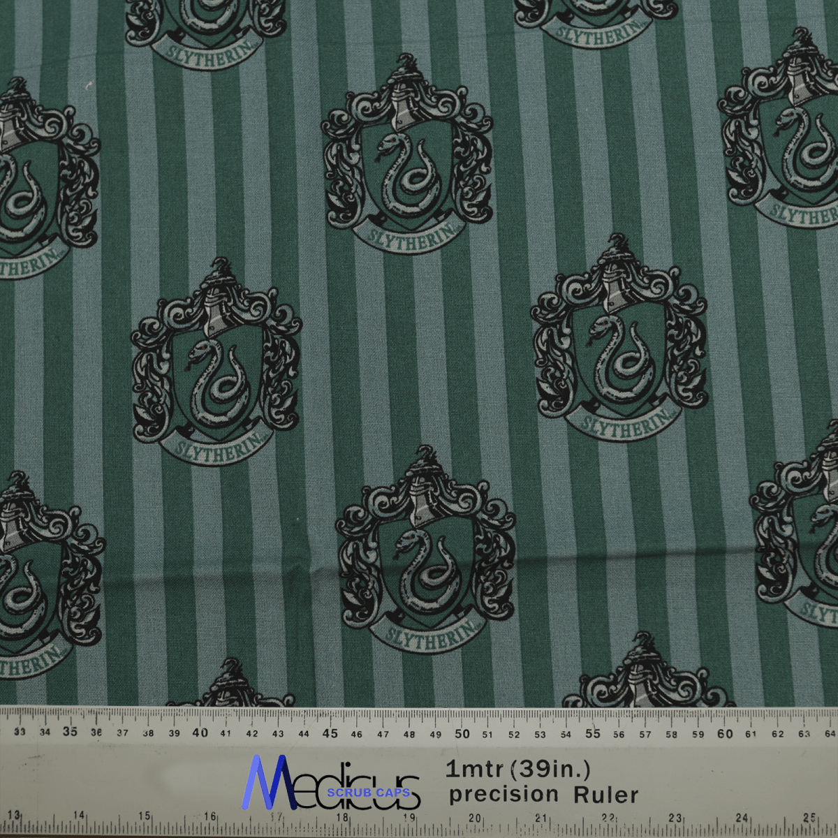 Green fabric with vertical stripes displays Slytherin crests and custom embroidery, each featuring a serpent and decorative elements. An eco-friendly ruler with metric/inch markings is seen at the bottom, ideal for designing the Harry Potter Slytherin Scrub Cap by Medicus Scrub Caps.