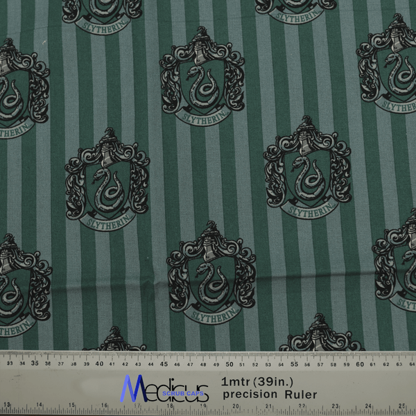 Green fabric with vertical stripes displays Slytherin crests and custom embroidery, each featuring a serpent and decorative elements. An eco-friendly ruler with metric/inch markings is seen at the bottom, ideal for designing the Harry Potter Slytherin Scrub Cap by Medicus Scrub Caps.