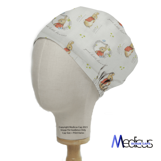 A mannequin models the Peter Rabbit Grey Cartoon Scrub Cap by Medicus Scrub Caps, showcasing charming Beatrix Potters Peter Rabbit with delicate flowers on a light background. The logo appears at the bottom right, adding style and nostalgia to this whimsical accessory.