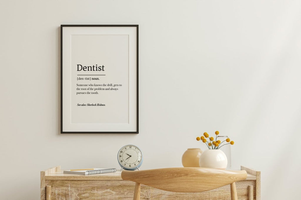 A digital download of the Dentist Definition Poster from Medicus Scrub Caps adorns the wall. Below, a wooden desk with a white clock, yellow-flowered vase, and book beside a wooden chair perfectly complements your Medicus Scrub Caps.