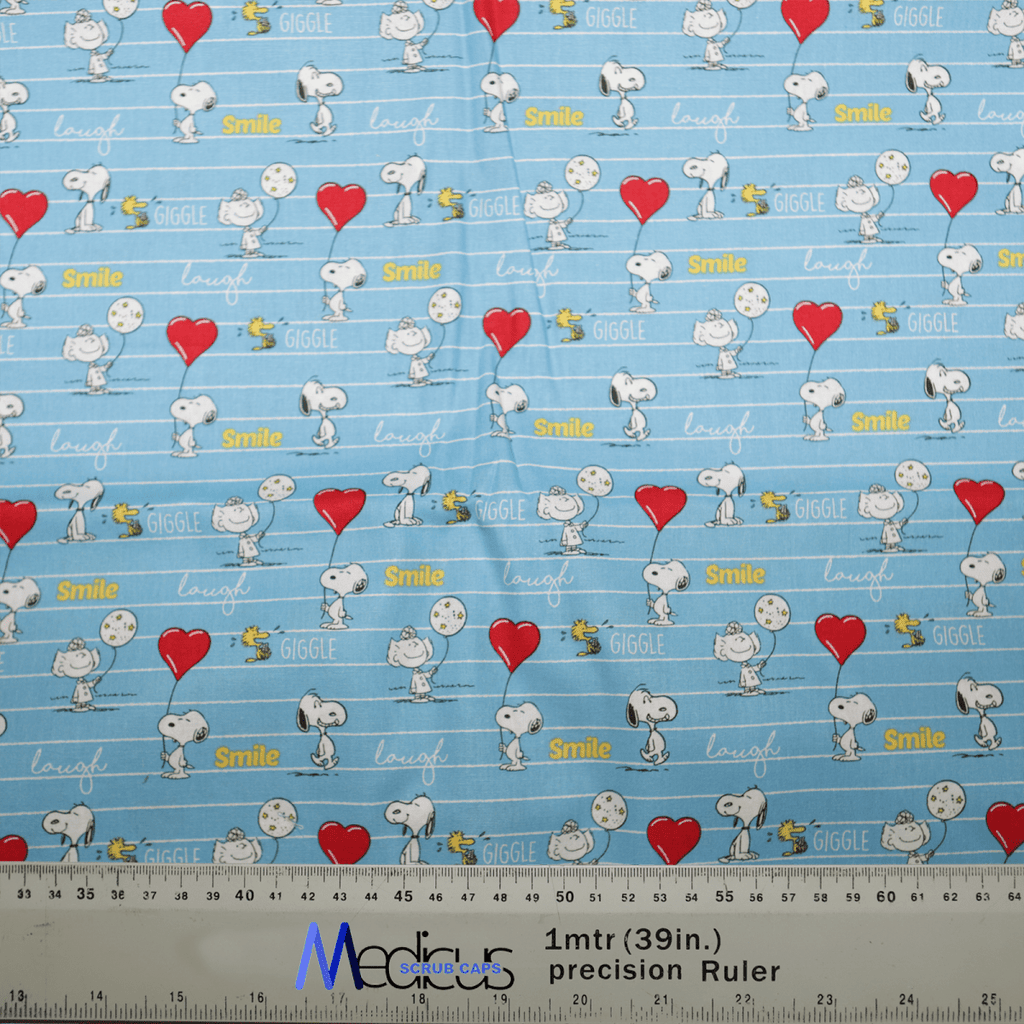 The Medicus Scrub Caps Snoopy Heart Balloon Laugh Love Scrub Cap delights with cartoon dogs, birds, hearts, and words like Giggle, Smile, and Laugh on a blue background; a ruler in inches and centimeters provides scale.