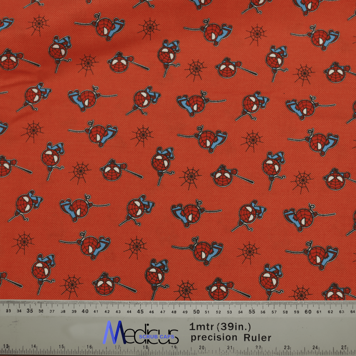 The Spiderman Kawaii On Red Scrub Cap from Medicus Scrub Caps features a vibrant red fabric with cartoon-style masked faces, spider webs, and spiders, ideal for crafting superhero scrub hats. A gray ruler at the bottom offers measurements in meters and inches for precise cutting.