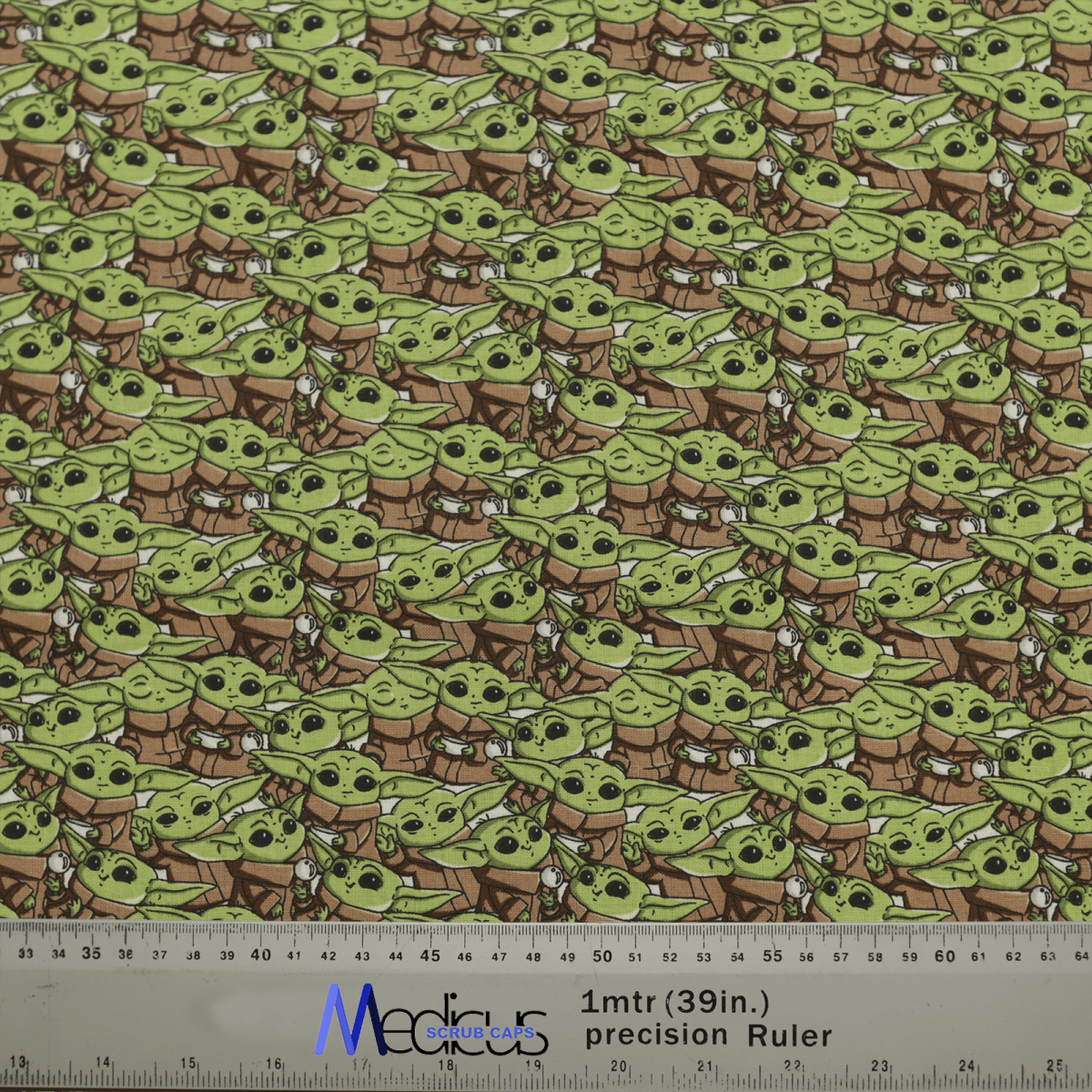 Patterned fabric with a small green alien in a brown robe and white scarf adds intergalactic charm. A 1-meter precision ruler shown for scale. Ideal for making a Star Wars Baby Yoda Crowd Scrub Cap by Medicus Scrub Caps with style.